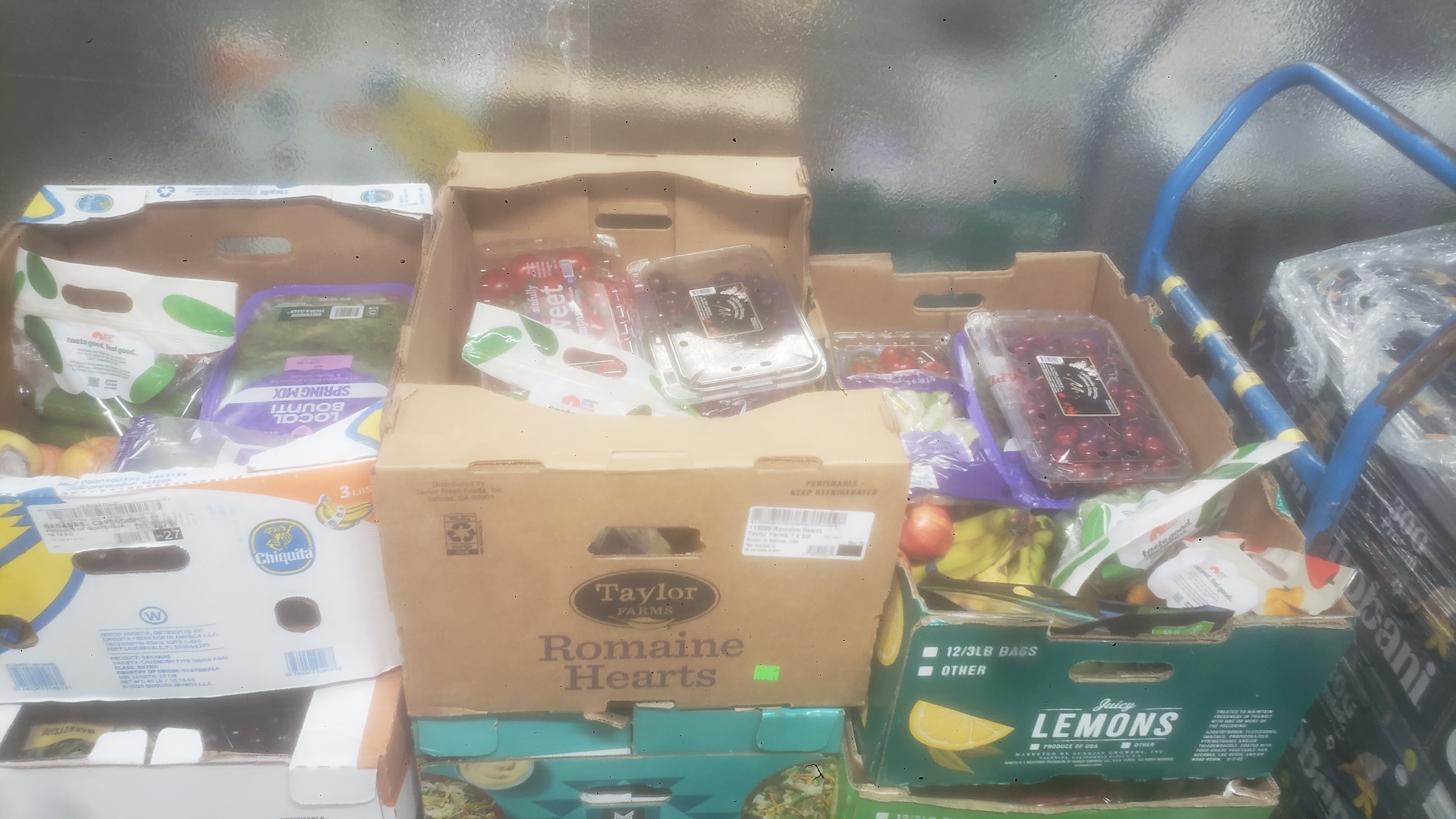 Stacks of produce boxes filled with produce such as a container of cherries, apples, and a bag of cucumbers.